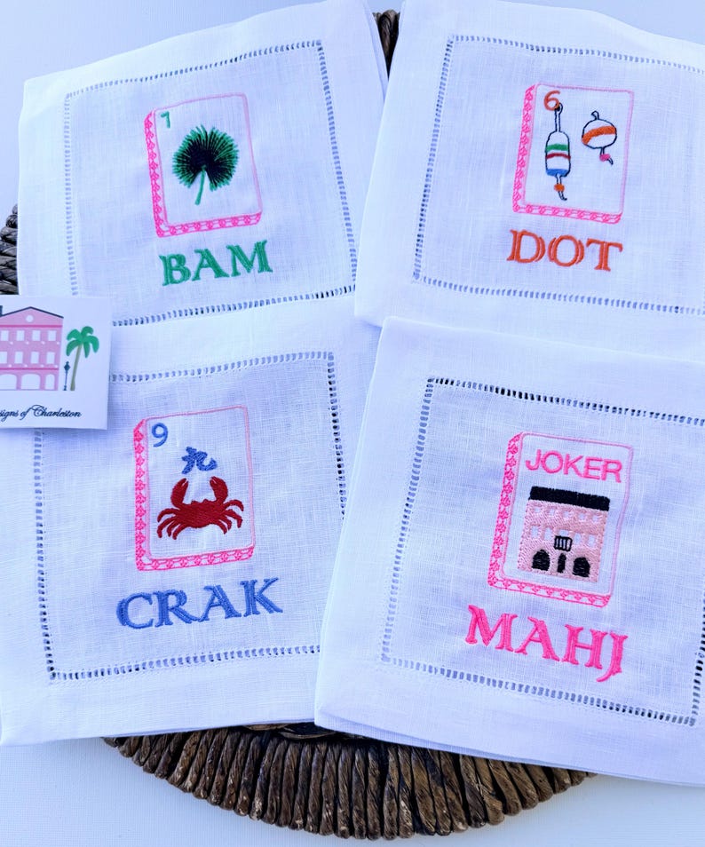 Może przedstawiać: Cztery kwadratowe białe lniane serwetki z wyhaftowanymi wzorami płytek mahjong. Każda serwetka przedstawia unikalną grafikę z napisami BAM, DOT, CRAK i MAHJ, wykonanymi przy użyciu żywych kolor&oacute;w, takich jak zielony, pomarańczowy, niebieski i r&oacute;żowy.