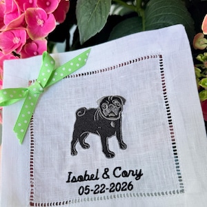 May include: A square white linen napkin featuring an embroidered black pug dog, the names "Isabel & Cory", and the date "05-22-2026" in black thread. The napkin has a decorative hemstitch border and a light green ribbon with white polka dots.