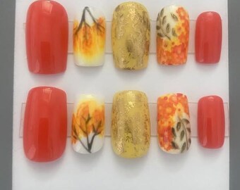 Peut inclure: Un ensemble de dix faux ongles aux designs variés. Les ongles comprennent du rouge uni, du papier doré et des motifs d'automne aux couleurs orange, jaune et blanc. Les ongles sont présentés sur une surface blanche.