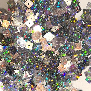 May include: A pile of iridescent silver square sequins with a holographic finish. The sequins have a small hole in the center.