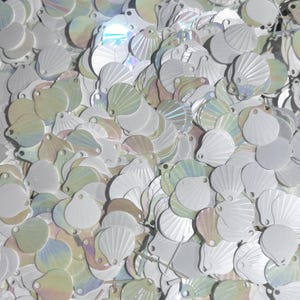 May include: A close-up shot of a pile of white, iridescent seashell-shaped sequins. The sequins have a pearlescent sheen, reflecting light with hints of pink, green, and blue. The sequins are scattered, creating a textured surface.