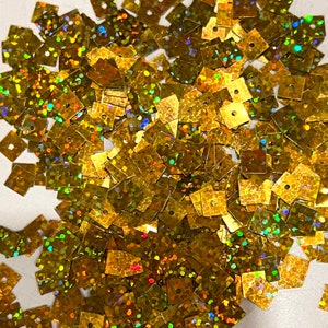 May include: A pile of gold square sequins with a holographic finish. The sequins have a small hole in the center.