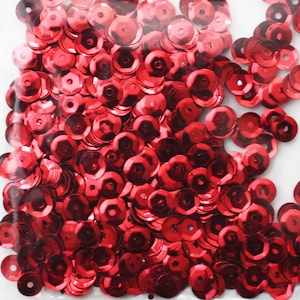 May include: A close-up image of a bag filled with red sequins. The sequins are small and round, with a shiny finish.