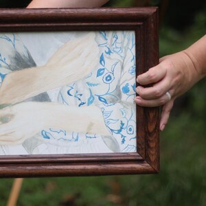 May include: A framed watercolour painting with a blue and white floral design. The artwork shows a pair of hands holding a light-coloured object. The frame is made of dark brown wood, giving the piece a classic look.