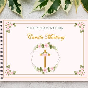 Puede incluir: Un cuaderno con espiral y cubierta rosa claro. El texto "MI PRIMERA COMUNIÓN" y "Camila Martínez" están en dorado. Una cruz dorada está centrada en una corona floral. Rosas rosas y hojas verdes decoran las esquinas.