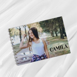May include: A spiral-bound photo album featuring a young person smiling outdoors. The album cover displays the name "CAMILA" in bold black letters, with "MIS 15 AÑOS" below. The background is a blurred outdoor scene.