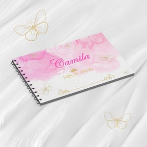 Puede incluir: Un cuaderno con espiral con un diseño de acuarela rosa y blanco. La portada presenta el nombre "Camila" en escritura rosa, una corona y el texto "MIS XV AÑOS". Acentos dorados de mariposas y flores completan el diseño.