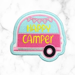 May include: A blue and pink embroidered patch with a camper van design. The camper van has a white roof and the words "Happy Camper" are embroidered in yellow on the side.