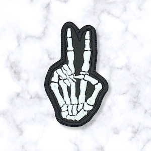 May include: A black embroidered patch with a white skeleton hand making the peace sign.