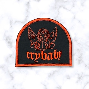 May include: Black iron-on patch with an orange outline. The patch features a cartoon angel with wings and the text "crybaby" in a gothic font.