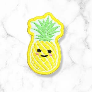 May include: A yellow embroidered pineapple with a green leafy top and a smiling face. The pineapple has a white outline and is on a white background.
