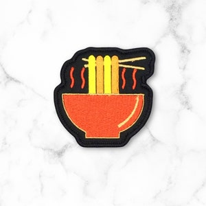May include: A black and red embroidered patch of a bowl of ramen noodles with chopsticks. The noodles are yellow and the bowl is red with a black outline.
