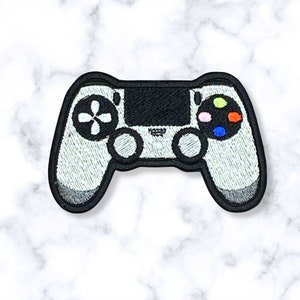 May include: A white and black embroidered patch of a video game controller. The controller has a black face with a white PlayStation logo and colorful buttons.