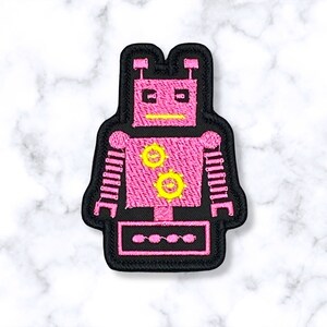 May include: Pink robot patch with yellow details. The robot has a round head with two antennas, two large eyes, and a mouth. The robot's body has two gears and two arms with stripes. The robot is standing on a black base with four circles.