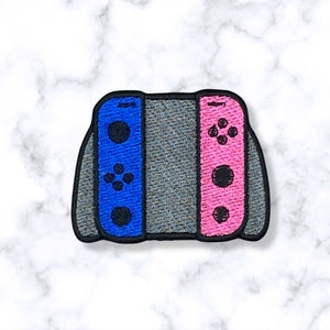 May include: A blue and pink embroidered patch of a Nintendo Switch console with the controllers attached.