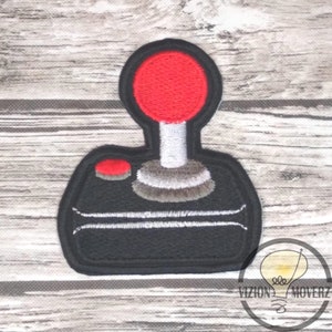 May include: A black and red embroidered patch of a vintage video game joystick. The joystick has a red button and a gray base.
