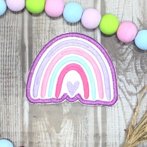 May include: A purple, pink, and blue rainbow with a small purple heart in the center. The rainbow is embroidered on a white fabric.