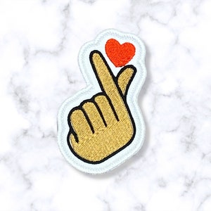 May include: White embroidered patch with a yellow hand making a heart shape with the index finger and thumb. A small red heart is on top of the index finger.