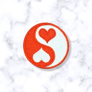 May include: A red and white yin yang symbol with two hearts, one red and one white, in the center. The symbol is embroidered on a circular patch.