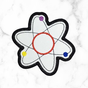 May include: A white embroidered patch with a black outline shaped like an atom. The atom has a red circle in the center and four colored dots around it: purple, yellow, blue, and blue.