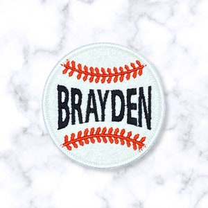 May include: White baseball-themed patch with red stitching and the name "Brayden" embroidered in black.