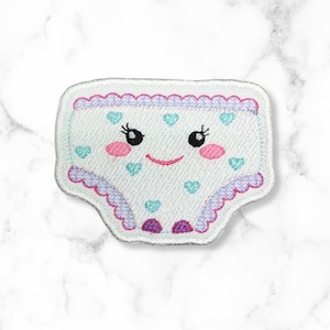 May include: White embroidered patch of a pair of underwear with a smiling face, pink cheeks, and blue hearts. The underwear has a purple and white scalloped trim.