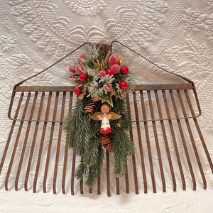 May include: A rusty metal rake decorated with a Christmas-themed garland. The garland features pine branches, red berries, white faux snow, and a small wooden angel figurine.