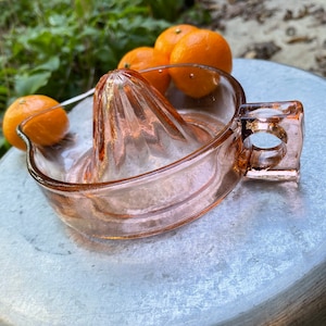 K&ouml;nnte beinhalten: Ein vintage pinkfarbener Glas-Zitronenpresse mit Griff. Die Presse steht auf einer silbernen Oberfl&auml;che mit vier Orangen in der N&auml;he.