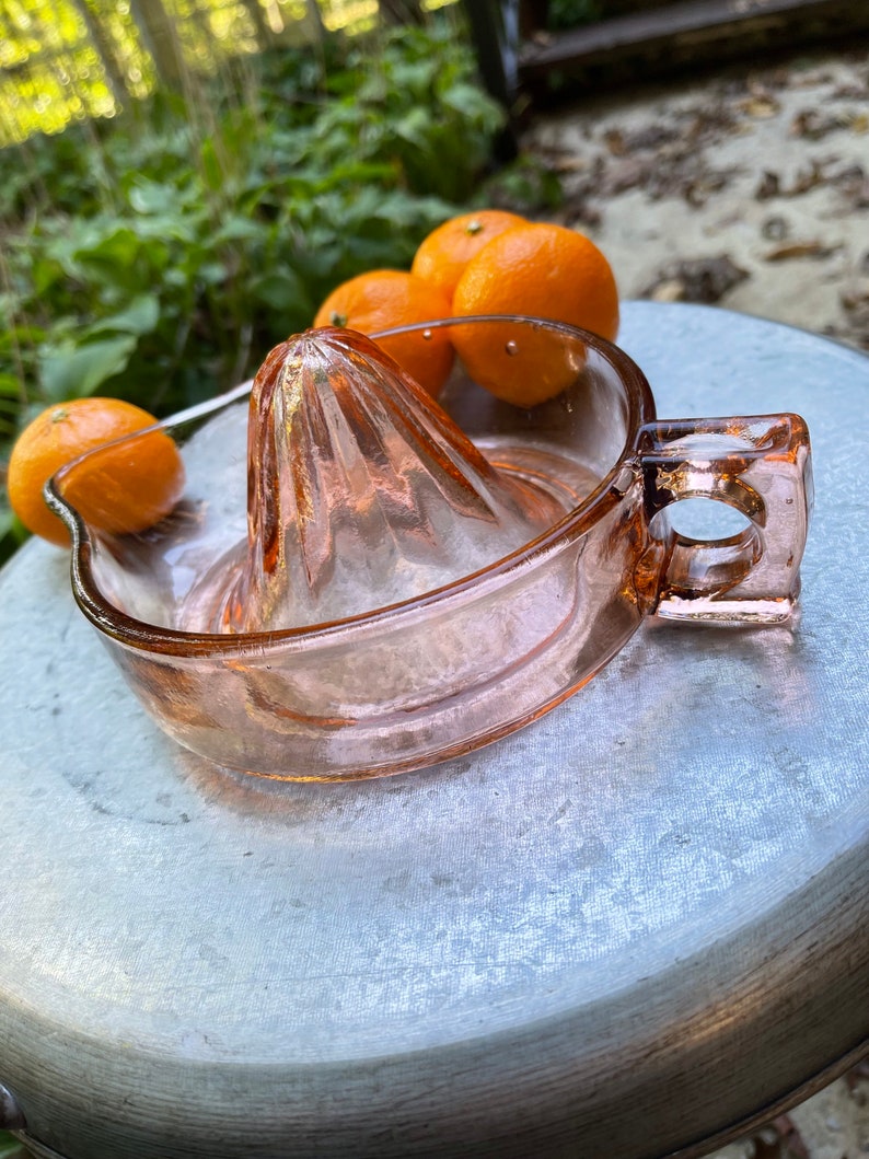 K&ouml;nnte beinhalten: Ein vintage pinkfarbener Glas-Zitronenpresse mit Griff. Die Presse steht auf einer silbernen Oberfl&auml;che mit vier Orangen in der N&auml;he.