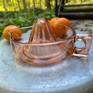K&ouml;nnte beinhalten: Ein rosa gl&auml;serner Zitronenpresse mit geripptem Design. Die Presse steht auf einer silbernen Oberfl&auml;che mit zwei Orangen im Hintergrund.