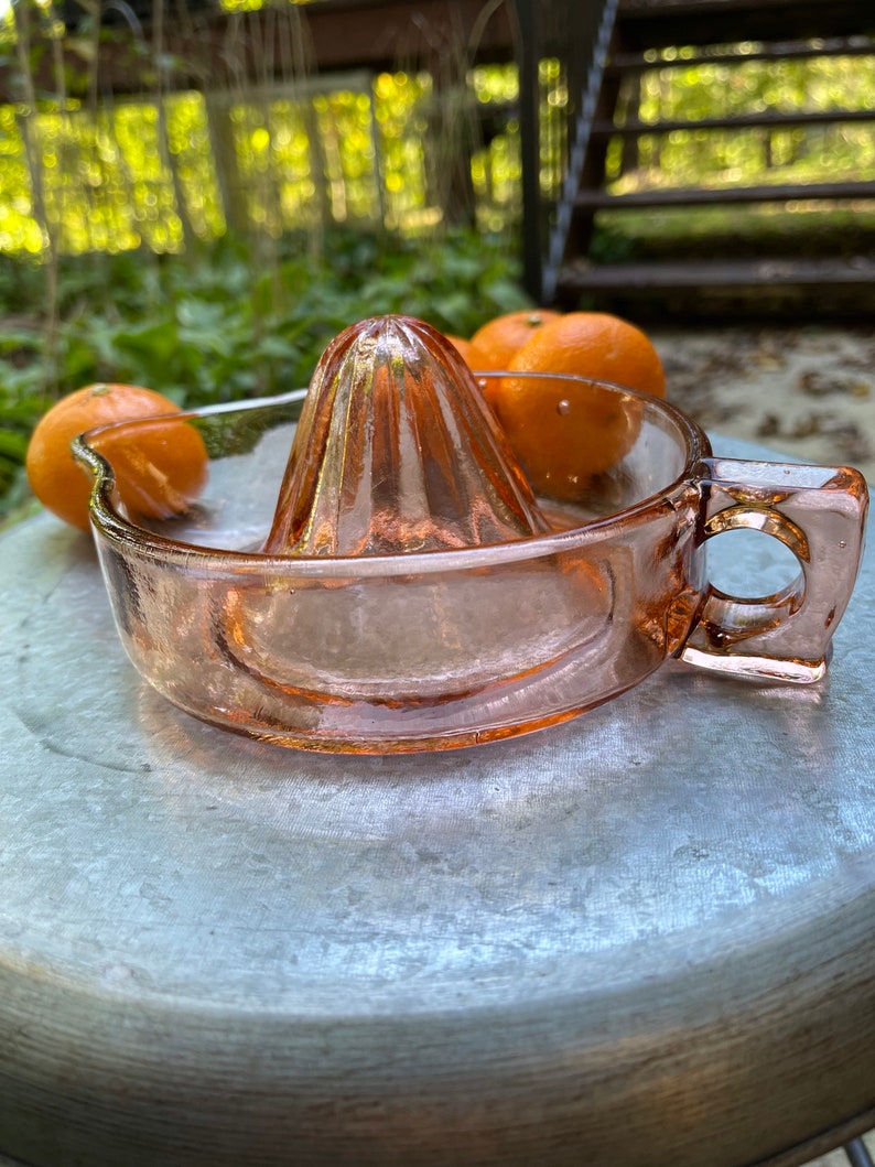 K&ouml;nnte beinhalten: Ein rosa gl&auml;serner Zitronenpresse mit geripptem Design. Die Presse steht auf einer silbernen Oberfl&auml;che mit zwei Orangen im Hintergrund.