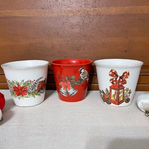 May include: Three ceramic flower pots with a Christmas theme. The pots are white with red and green designs. One pot has a red base and features a bird and bells. Another pot has a red bow and a lantern. The third pot has a poinsettia and pine cones.