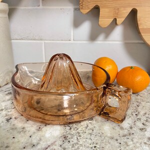 K&ouml;nnte beinhalten: Ein vintage Orangenpresse aus Glas mit einem Griff. Die Presse ist hellrosa und hat ein geriffeltes Design. Sie steht auf einer wei&szlig;en Arbeitsplatte mit zwei Orangen daneben.