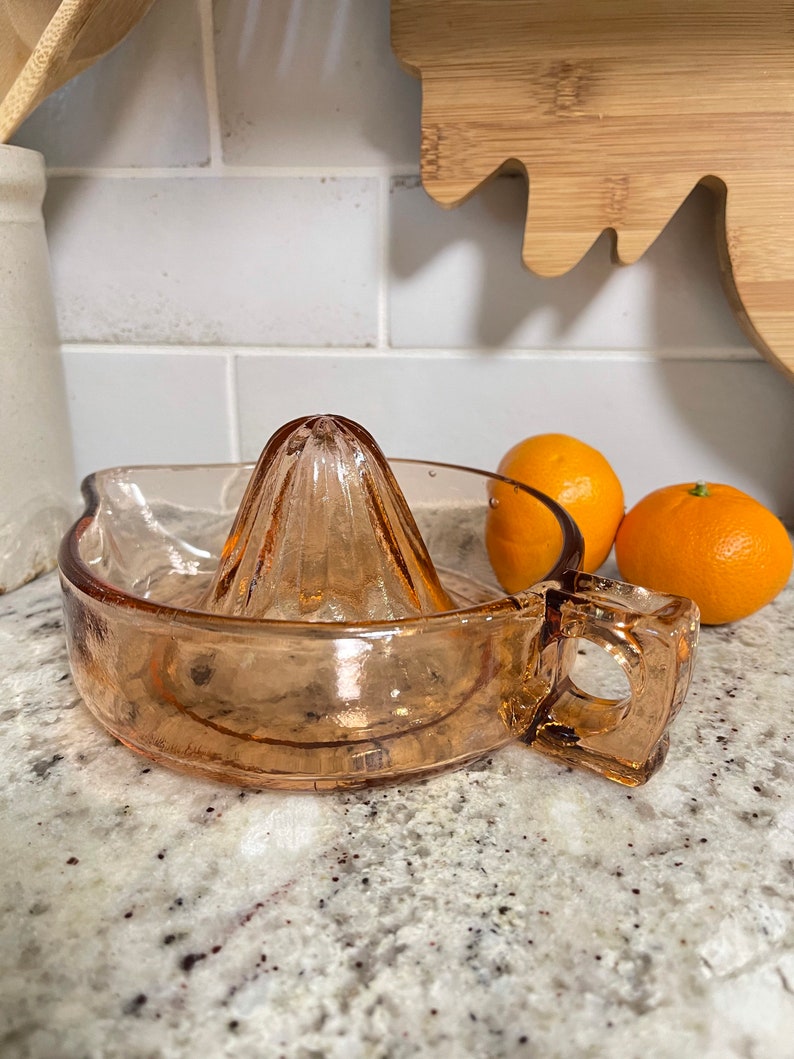 K&ouml;nnte beinhalten: Ein vintage Orangenpresse aus Glas mit einem Griff. Die Presse ist hellrosa und hat ein geriffeltes Design. Sie steht auf einer wei&szlig;en Arbeitsplatte mit zwei Orangen daneben.