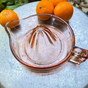 K&ouml;nnte beinhalten: Ein rosa gl&auml;serner Zitruspresse mit geriffeltem Zentrum und einem Griff. Vier ganze Orangen liegen auf einer silbernen Oberfl&auml;che hinter der Presse.
