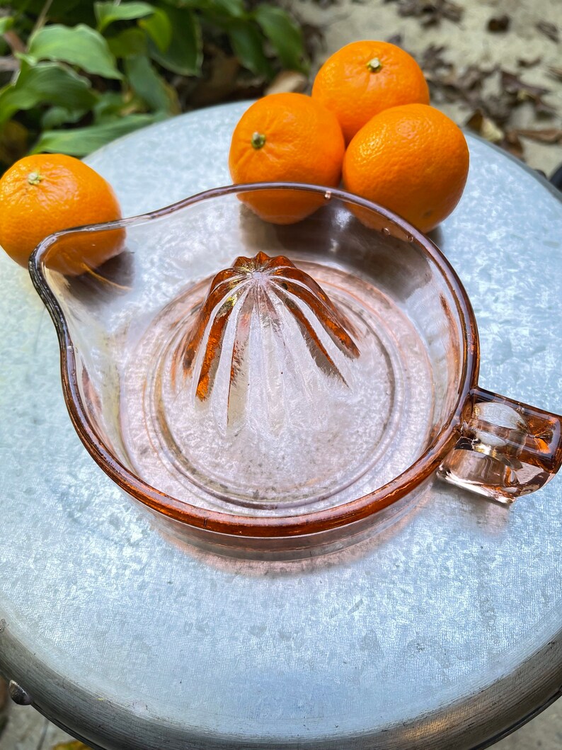 K&ouml;nnte beinhalten: Ein rosa gl&auml;serner Zitruspresse mit geriffeltem Zentrum und einem Griff. Vier ganze Orangen liegen auf einer silbernen Oberfl&auml;che hinter der Presse.