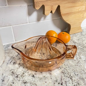 K&ouml;nnte beinhalten: Ein vintage Zitruspresse aus bernsteinfarbenem Glas mit geriffeltem Design. Die Presse hat einen Ausguss und einen Griff. Im Hintergrund befinden sich zwei Orangen.
