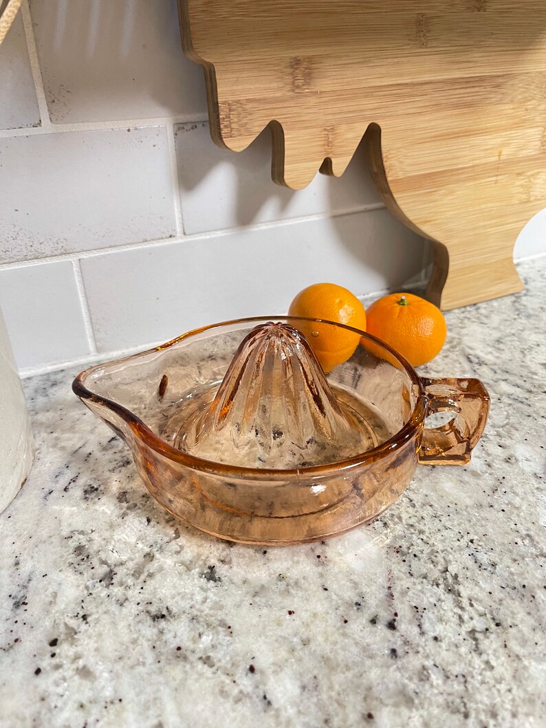 K&ouml;nnte beinhalten: Ein vintage Zitruspresse aus bernsteinfarbenem Glas mit geriffeltem Design. Die Presse hat einen Ausguss und einen Griff. Im Hintergrund befinden sich zwei Orangen.