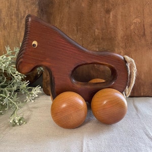 May include: A wooden toy horse with brown and natural wood tones. The horse has a pull string and four wheels.
