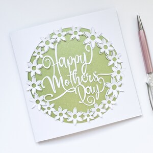 May include: A white card with a green circle in the center. The circle is surrounded by a white paper cut-out design of flowers. The text "Happy Mother's Day" is written in white inside the circle.