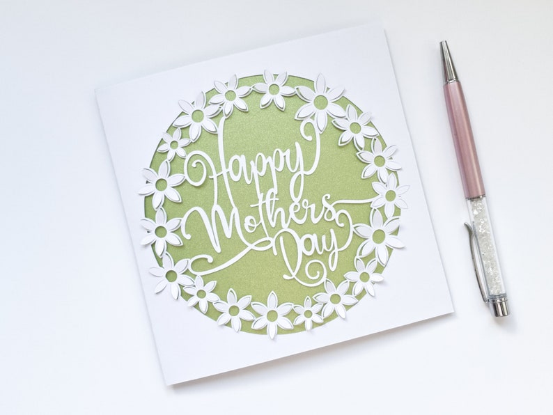 May include: A white card with a green circle in the center. The circle is surrounded by a white paper cut-out design of flowers. The text "Happy Mother's Day" is written in white inside the circle.