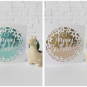 K&ouml;nnte beinhalten: Zwei wei&szlig;e Karten mit einem Kreis, der aus der Mitte ausgeschnitten ist. Die Kreise sind mit einem metallischen Hintergrund und wei&szlig;en Papierblumen gef&uuml;llt. Der Text "Happy Birthday" ist in Wei&szlig; auf dem metallischen Hintergrund geschrieben. Eine Karte hat einen t&uuml;rkisfarbenen metallischen Hintergrund und die andere einen goldenen metallischen Hintergrund.