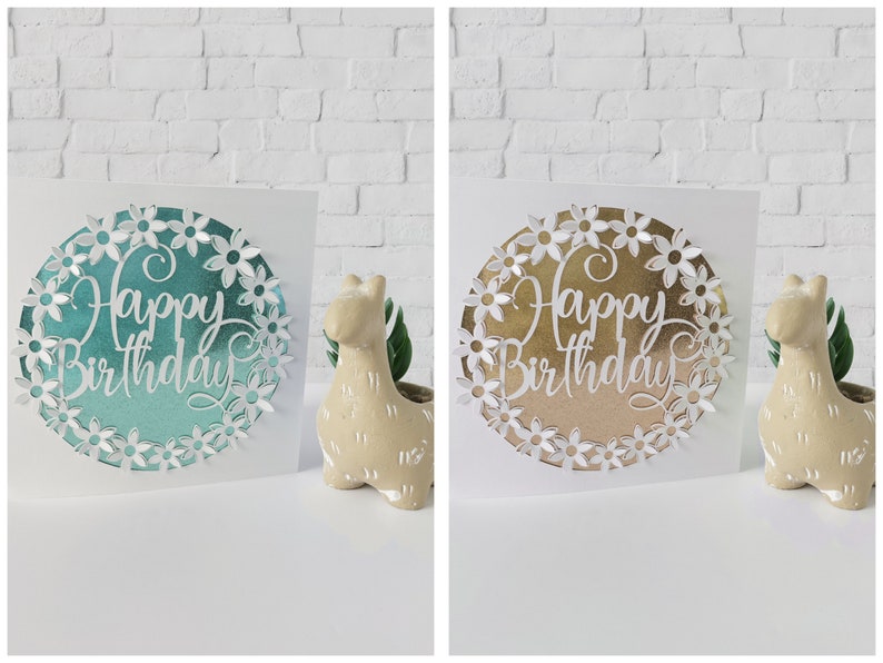 K&ouml;nnte beinhalten: Zwei wei&szlig;e Karten mit einem Kreis, der aus der Mitte ausgeschnitten ist. Die Kreise sind mit einem metallischen Hintergrund und wei&szlig;en Papierblumen gef&uuml;llt. Der Text "Happy Birthday" ist in Wei&szlig; auf dem metallischen Hintergrund geschrieben. Eine Karte hat einen t&uuml;rkisfarbenen metallischen Hintergrund und die andere einen goldenen metallischen Hintergrund.