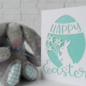 Puede incluir: Una tarjeta blanca con un huevo de purpurina turquesa recortado. El huevo tiene un conejito blanco sosteniendo flores y las palabras "Feliz Pascua" recortadas. Un conejito de peluche gris se sienta frente a la tarjeta.