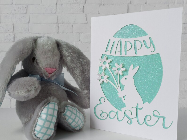 Puede incluir: Una tarjeta blanca con un huevo de purpurina turquesa recortado. El huevo tiene un conejito blanco sosteniendo flores y las palabras "Feliz Pascua" recortadas. Un conejito de peluche gris se sienta frente a la tarjeta.