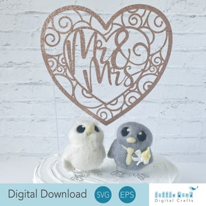 Op de afbeelding: Een hartvormige uitsnijding van roze glitter met de woorden "Mr & Mrs" erin. Twee kleine viltvogels, een wit en een grijs, staan op een witte taartstandaard.
