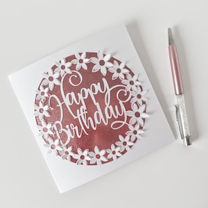 K&ouml;nnte beinhalten: Eine wei&szlig;e Karte mit einem ros&eacute;goldenen Kreis in der Mitte. Der Kreis hat ein wei&szlig;es Papier-Cut-Out-Design aus Blumen um die Worte "Happy Birthday".