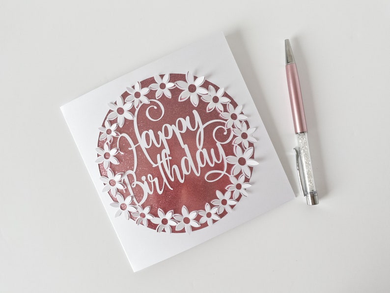 K&ouml;nnte beinhalten: Eine wei&szlig;e Karte mit einem ros&eacute;goldenen Kreis in der Mitte. Der Kreis hat ein wei&szlig;es Papier-Cut-Out-Design aus Blumen um die Worte "Happy Birthday".