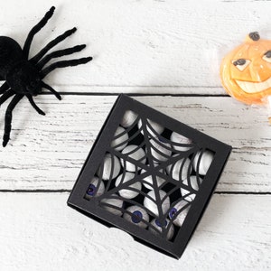 Pu&ograve; includere: Una scatola nera con un design a ragnatela tagliato riempita di occhi di caramelle bianchi. Un ragno nero e un lecca-lecca a forma di zucca arancione sono su una superficie di legno bianco.