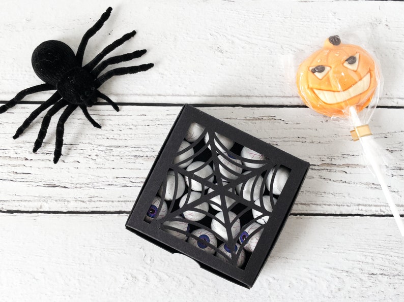 Pu&ograve; includere: Una scatola nera con un design a ragnatela tagliato riempita di occhi di caramelle bianchi. Un ragno nero e un lecca-lecca a forma di zucca arancione sono su una superficie di legno bianco.